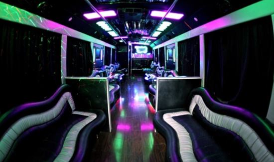 Party bus interior with curved black and white seating, glossy wood floors, and vibrant purple and green lighting.