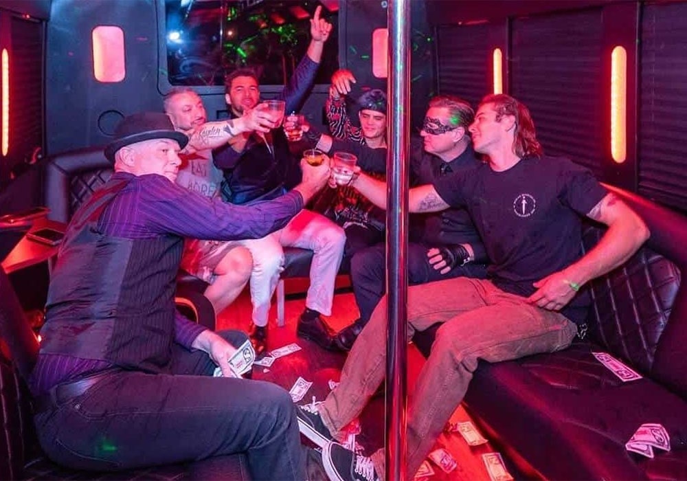 Group of men on a bachelor party bus brewery tour, raising drinks in a toast amid red neon lights.