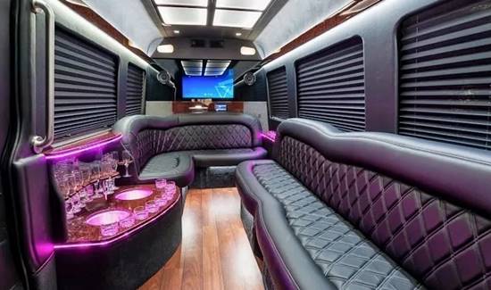 Party bus interior with black leather seating, pink accent lighting, wood floors, and a built-in drink station.