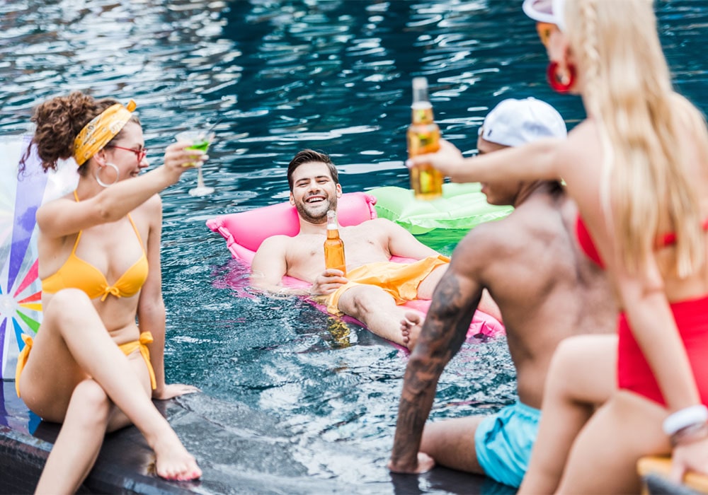 Group of young adults laughing and toasting with beer and cocktails by the pool.