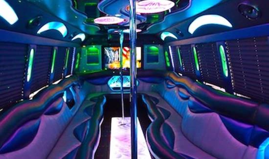 Interior of a luxury party bus with curved leather seating on both sides, neon lighting, and dance poles.