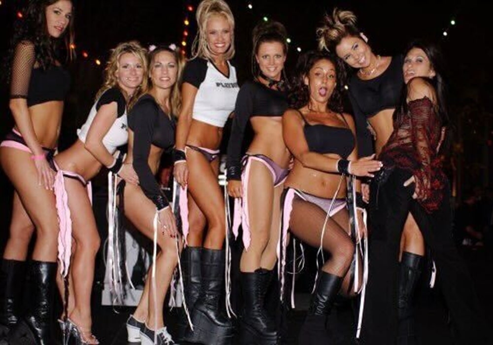 Liz with six Bad Girl entertainers posing in PlayboyTV-branded outfits at a lively nighttime party.
