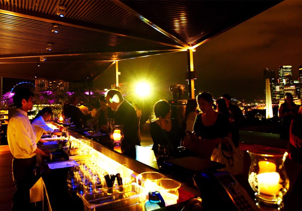 Bartenders and people at a rooftop bar by night with city lights in the background.