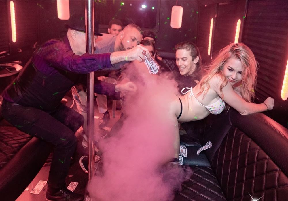 A young blonde stripper dances amid smoke on a bachelor party bus, receiving tips in her bikini.