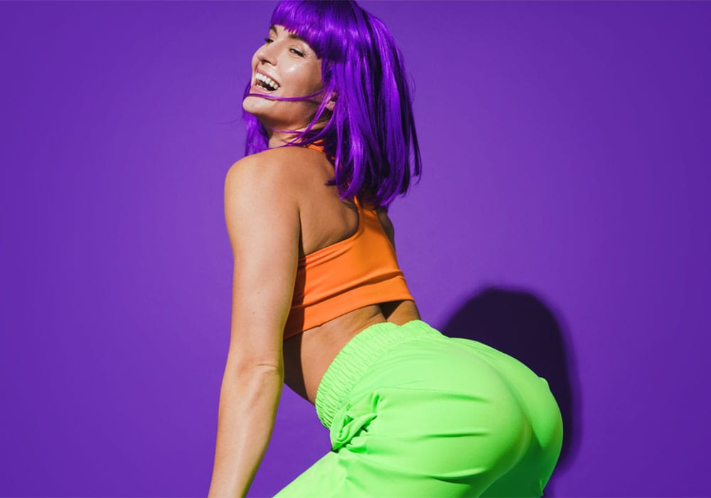 Sexy woman wearing colorful outfit twerking against purple background.