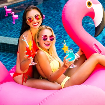 Two smiling young women in bikinis with cocktails sitting on a pink flamingo float in a swimming pool.