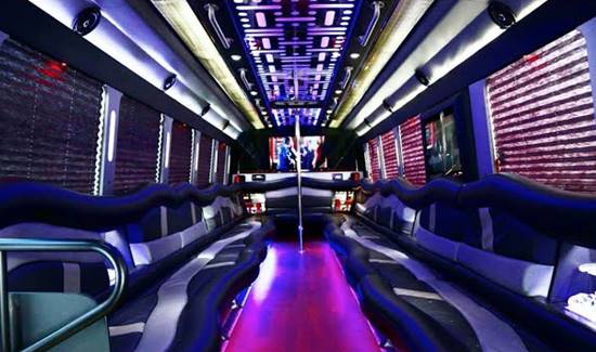 Interior of a luxury party bus with long leather couches along both sides, purple neon lighting, and a dance pole.