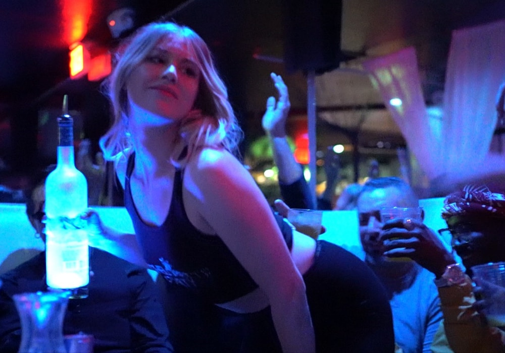 A young blonde female bartender dancing for a party group while holding a bottle of alcohol.